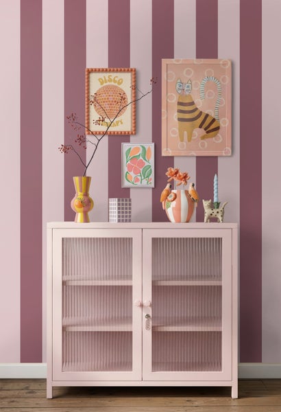 Hellrosa Metallschrank mit geriffelten Glastüren vor einer rosa gestreiften Wand, dekoriert mit bunten Vasen und gerahmten Bildern.