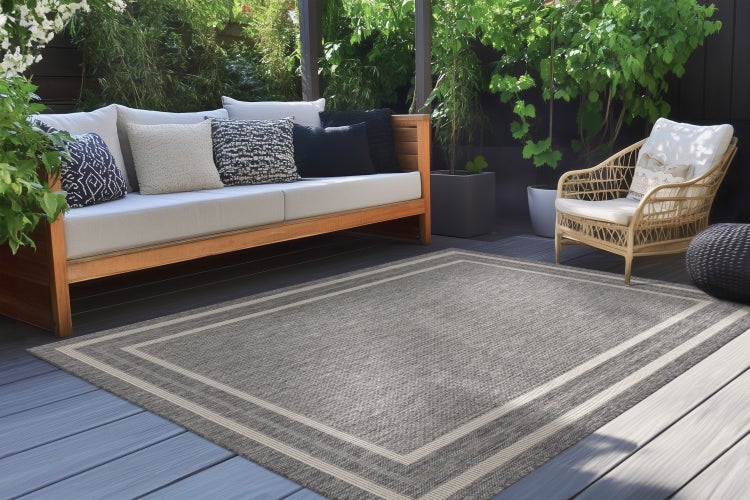 Rechteckiger Outdoor Teppich mit Bordürenmuster auf einer Holzterrasse vor einem Gartensofa und einem Rattan Sessel.