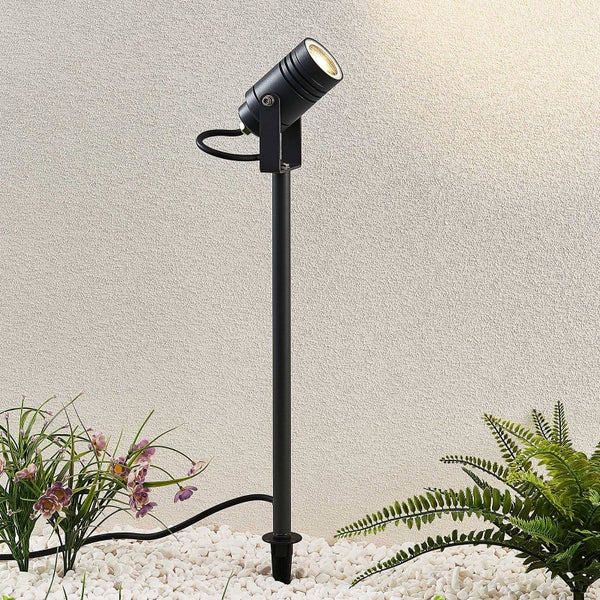 Verstellbarer LED-Gartenstrahler mit Erdspieß in einem Beet mit Pflanzen und Kies vor einer beleuchteten Wand.