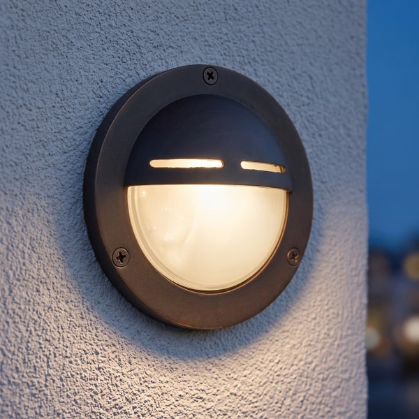 Runde LED Wandleuchte für den Außenbereich mit Schirm zur Lichtlenkung an einer hellen Fassade.