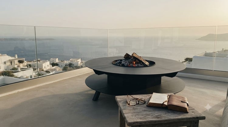 Runder Feuerring Grill mit Holzablage auf einer Terrasse mit Meerblick. Im Vordergrund ein Holztisch mit Buch und Brille.