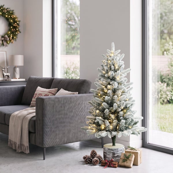 Ein künstlicher, verschneiter Weihnachtsbaum mit LED Beleuchtung in einem modern eingerichteten Wohnzimmer neben einem Sofa.