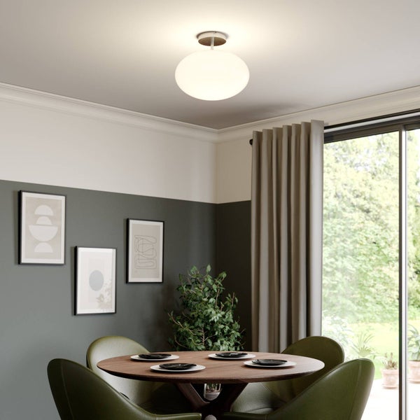 Moderne Deckenleuchte mit ovalem Glasschirm über einem runden Holztisch mit grünen Stühlen in einem hellen Esszimmer.