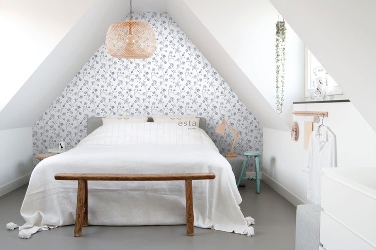 Hell eingerichtetes Schlafzimmer unter einer Dachschräge mit floraler Mustertapete, hellem Bett und einer Flechtlampe.