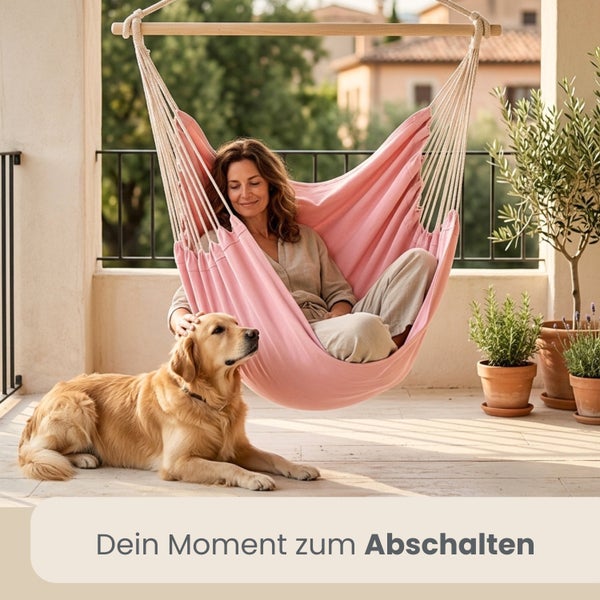 Frau sitzt in einem rosa Hängesessel aus Stoff mit Holzstab auf einem hellen Balkon neben einem Golden Retriever und Topfpflanzen.