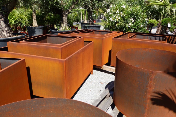 Quadratische und runde Pflanzgefäße aus Cortenstahl in einem hellen Gartencenter mit Bäumen und Sträuchern im Hintergrund.