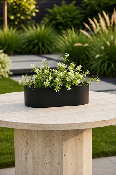 Ovales schwarzes Pflanzgefäß mit weißen Blüten auf einem Steintisch im Garten.