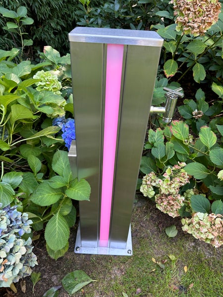 Wasserzapfstelle aus Edelstahl mit integrierter LED-Beleuchtung und Wasserhahn in einem Garten mit Hortensien.