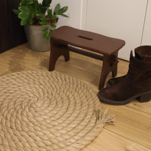 Ein runder Teppich aus dickem, gewundenem Seil liegt auf einem Holzboden neben einem kleinen Holzhocker, braunen Stiefeln und einer Topfpflanze.