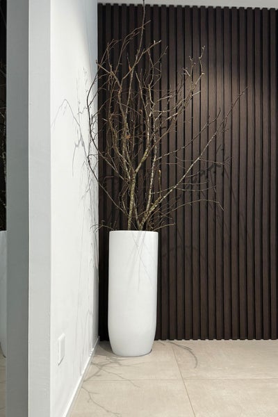 Hoher, weißer zylindrischer Pflanzkübel mit dekorativen Ästen vor einer Wand aus dunklen Holzlamellen und einer weißen Wand.