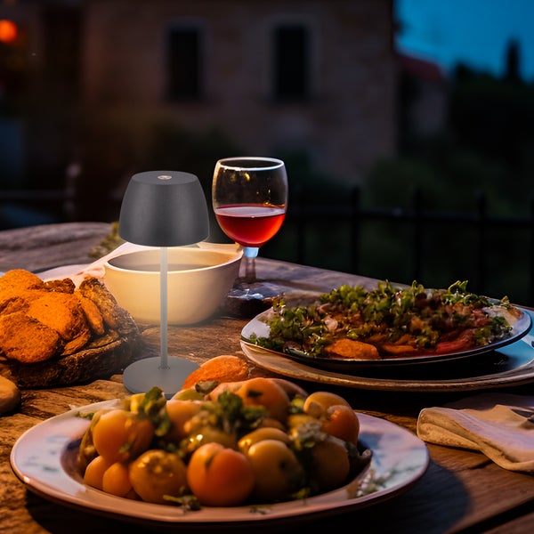 Schwarze LED-Tischleuchte auf einem hölzernen Tisch im Freien mit Speisen und Wein in abendlicher Atmosphäre.