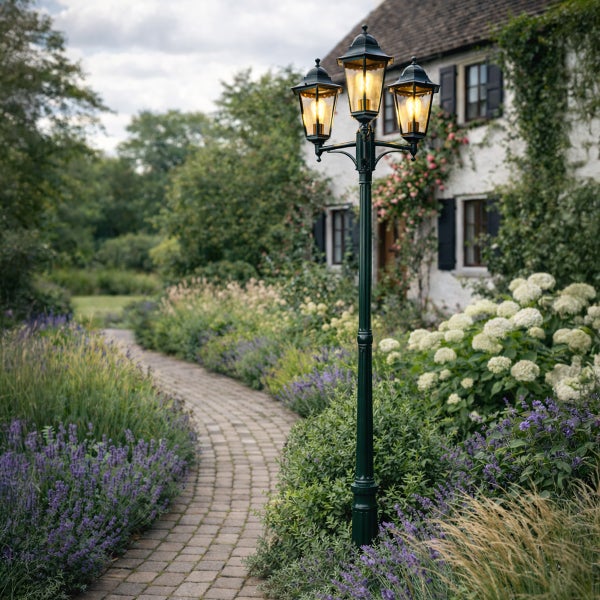 Dreiflammige Gartenlaterne im klassischen Design an einem gepflasterten Weg in einem blühenden Garten vor einem Wohnhaus.