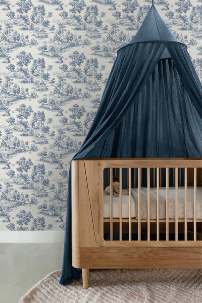Kinderzimmer mit Toile-de-Jouy-Tapete mit blauen Landschaftsmustern, einem hellen Babybett aus Holz und einem dunkelblauen Stoffhimmel.
