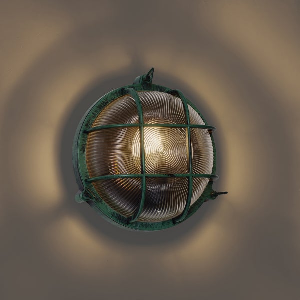 Runde Außenwandleuchte im nautischen Stil aus grün patiniertem Metall mit Schutzgitter und geripptem Glas, eingeschaltet.