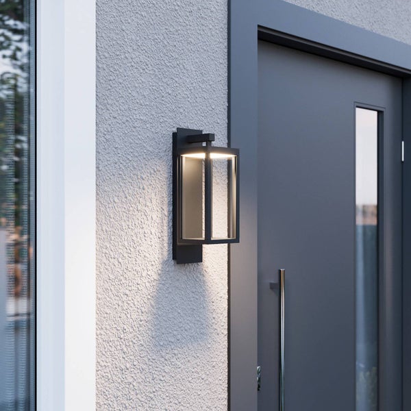 Moderne rechteckige Außenwandleuchte in Laternenform aus Metall mit Glaseinsätzen, montiert an einer strukturierten Hauswand neben einer Haustür.