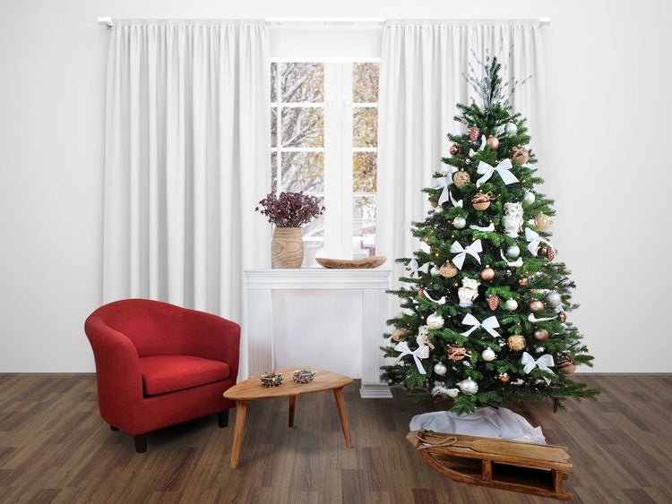 Festlich geschmückter Weihnachtsbaum mit Kugeln und Schleifen in einem hellen Wohnraum mit Polstersessel, Beistelltisch und Holzschlitten.