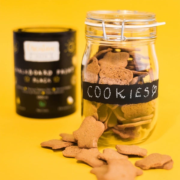 Ein Vorratsglas mit Keksen, versehen mit einem Streifen Tafelfarbe und der Aufschrift Cookies. Im Hintergrund steht eine Dose Tafelfarbe.
