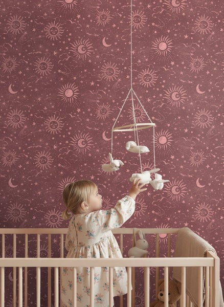 Kind im Holz-Gitterbett vor Tapete mit Sonnen-, Mond- und Sternenmuster, greift nach einem Baby-Mobile mit Stofftieren.