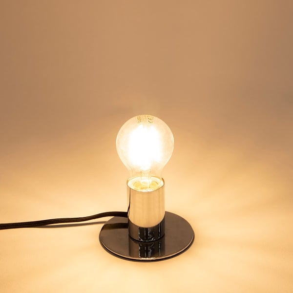 Tischleuchte mit schwarzem Fuß, verchromter Fassung und eingeschalteter Glühbirne.