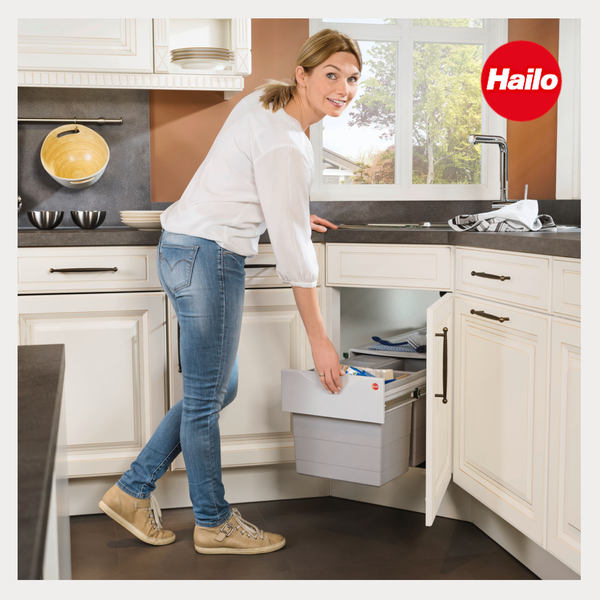 Eine Frau bedient ein ausziehbares Hailo Abfalltrennsystem in einem weißen Küchenunterschrank in einer hellen Küche. Hailo Logo.