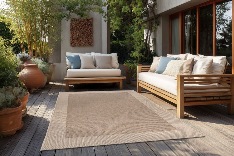 Ein rechteckiger Outdoor Teppich in gewebter Optik auf einer Holzterrasse zwischen zwei Gartensofas mit Kissen und zahlreichen Pflanzen.