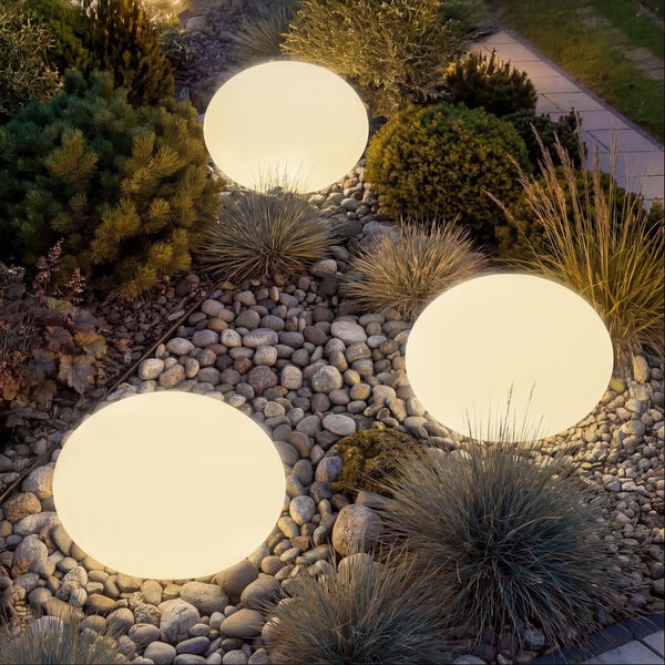 Drei leuchtende Garten-Kugelleuchten in einem Beet mit Kieselsteinen und verschiedenen Ziergräsern.