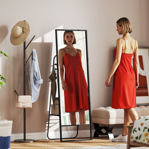 Innenraumszene mit einer jungen Frau, die ein rotes Kleid vor einem stehenden Spiegel betrachtet, ergänzt durch einen Kleiderständer und eine Bank
