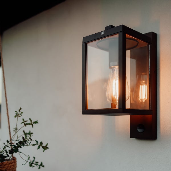 Außenwandleuchte mit schwarzem Metallrahmen, Glaseinsätzen, Bewegungsmelder und leuchtender Filamentlampe an einer hellen Fassade.