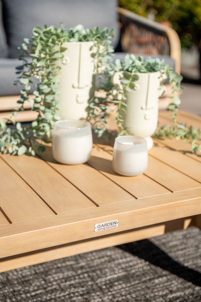 Garten-Couchtisch in Holzoptik mit Garden Impressions Logo, dekoriert mit Vasen in Gesichtsform, grünen Pflanzen und Kerzenhaltern.