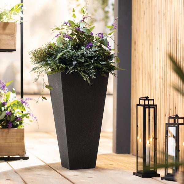 Hoher konischer Pflanzkübel in Anthrazit mit Blumen und Farnen auf einer Holzterrasse neben schwarzen Laternen vor einer Holzwand.