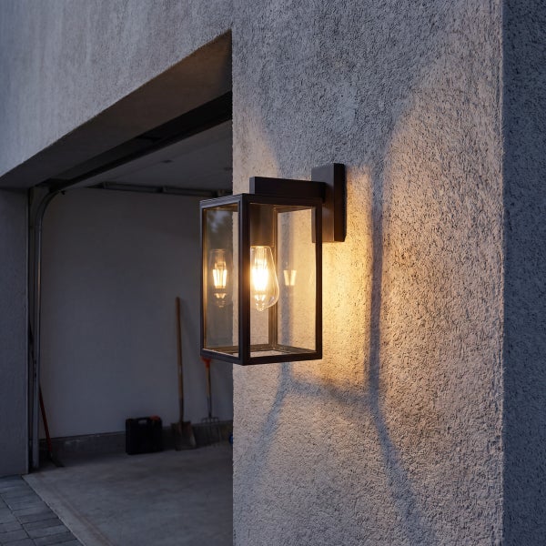 Moderne Außenwandleuchte in Laternenform mit Filament-Leuchtmittel an einer Hauswand neben einer Garage.