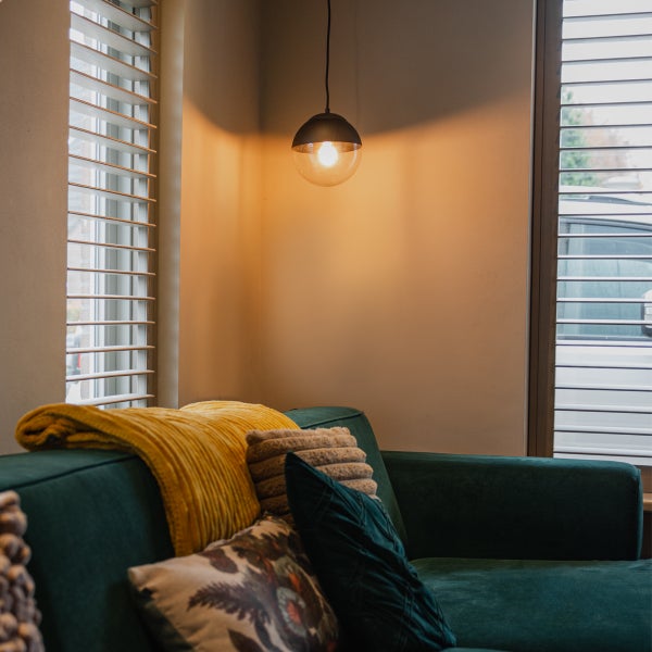 Wohnzimmer mit grüner Couch, Kissen, Plaid, Hängelampe und Fensterjalousien.