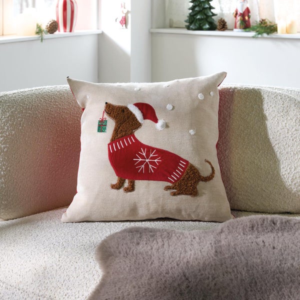 Quadratisches Weihnachtskissen mit Dackelmotiv, trägt Weihnachtsmütze und Pullover, liegt auf hellem Sofa, dekoratives Wohntextil für Weihnachten.