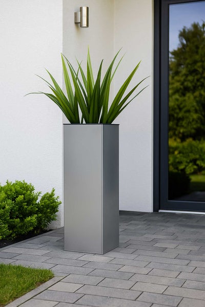 Hoher, rechteckiger Pflanzkübel in Silber-Optik mit einer grünen Pflanze auf einer gepflasterten Terrasse vor einer weißen Hauswand.