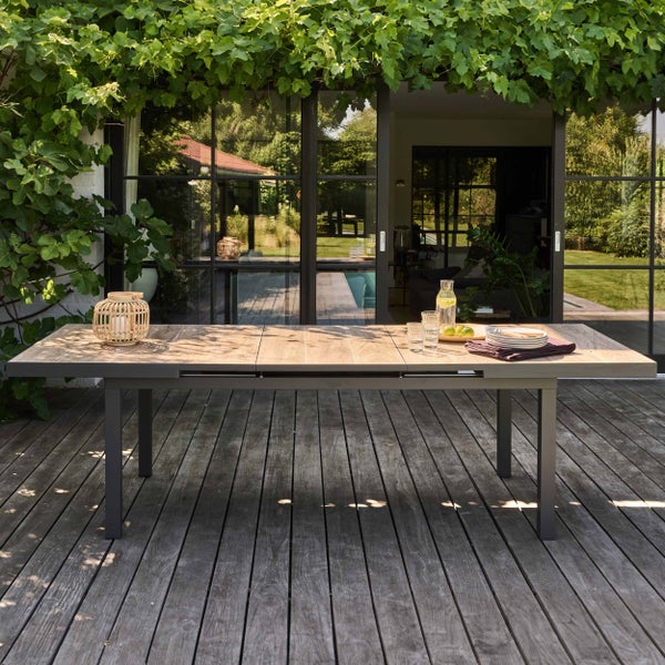 Ausziehbarer Gartentisch mit grauem Aluminiumgestell und Platte in Holzoptik auf einer Holzterrasse im hellen und organisierten Gartenbereich.
