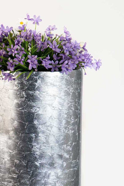 Hoher Pflanzkübel in Silber-Optik mit strukturierter Oberfläche und violetten Kunstblumen.