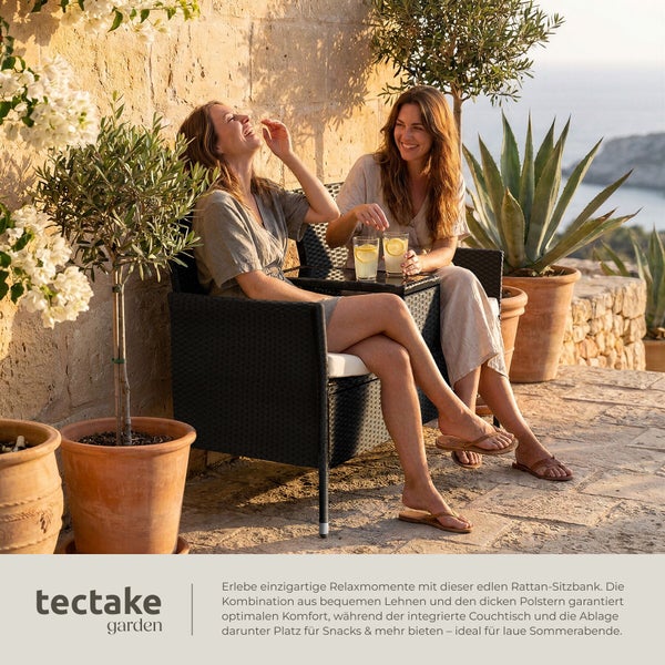 Zwei Frauen sitzen auf einer Rattan-Gartenbank mit integriertem Tisch auf einer sonnigen Steinterrasse mit Olivenbäumen in Töpfen.