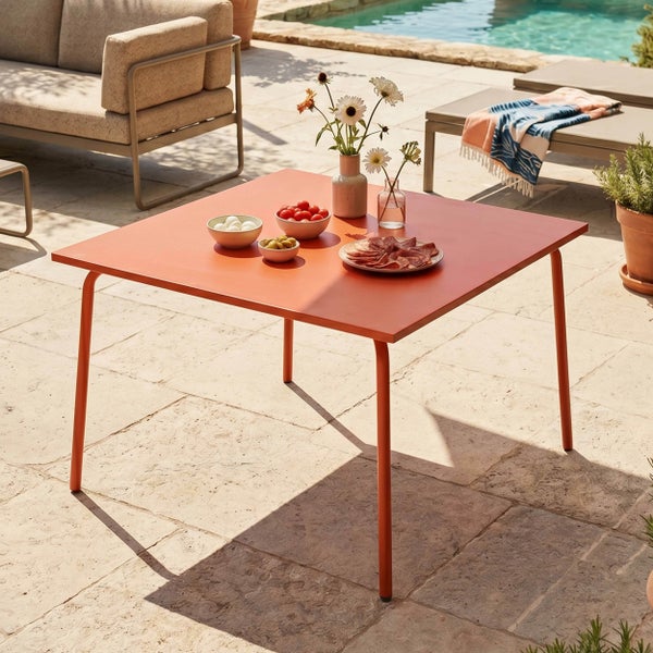 Quadratischer Gartentisch aus Metall auf einer hellen Steinterrasse mit Pool im Hintergrund, dekoriert mit Snacks und Blumenvasen.