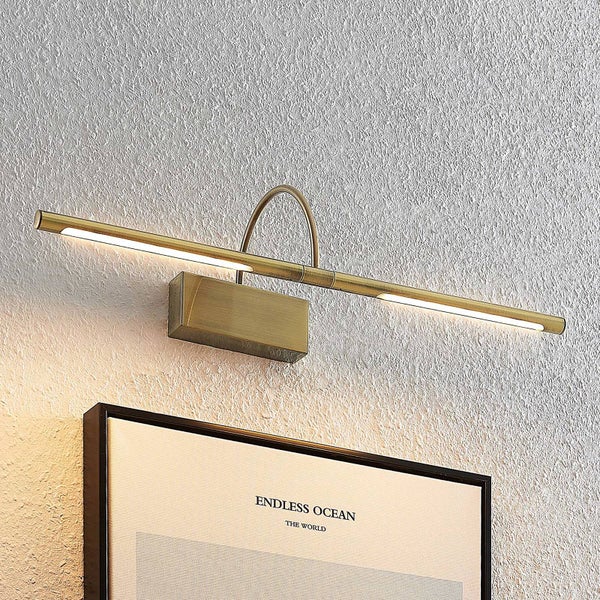 Moderne LED-Bilderleuchte in Messingoptik, montiert über einem gerahmten Bild an einer strukturierten Wand.