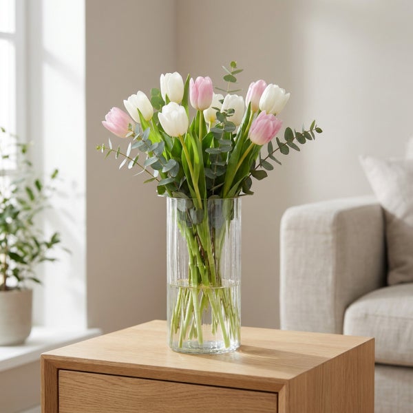 Strauß aus Tulpen und Eukalyptuszweigen in einer gerippten Glasvase auf einem Holztisch in einem Wohnzimmer.