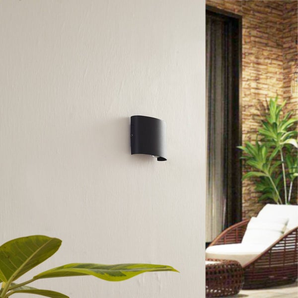 Moderne schwarze gebogene Außenwandleuchte montiert an einer hellen Wand auf einer Terrasse mit Liegestuhl und Pflanzen.