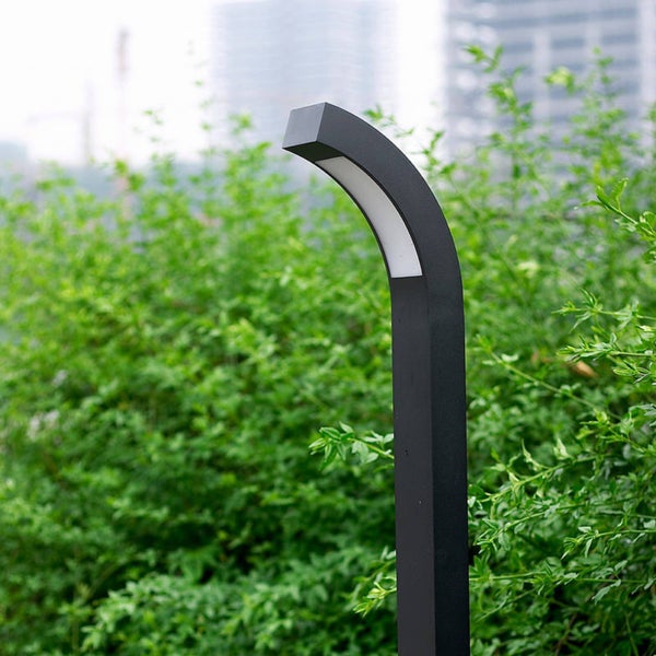 Moderne LED-Wegleuchte mit gebogenem Kopf aus anthrazitfarbenem Metall im Garten vor grünen Sträuchern.