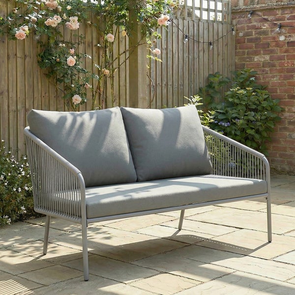 Graues Zweisitzer Gartensofa mit Polstern und Kordelbespannung auf einer Terrasse vor einem Holzzaun mit Rosen.