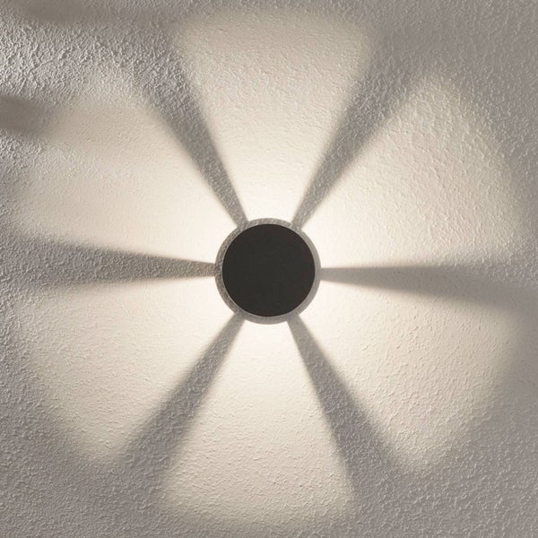 Schwarze runde Wandleuchte mit sechs Lichtstrahlen, die ein sternförmiges Muster auf eine strukturierte Wand werfen.