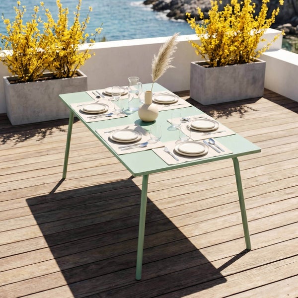 Moderner grüner Gartentisch für sechs Personen auf einer Holzterrasse mit Meerblick und Pflanzgefäßen mit gelben Blumen.