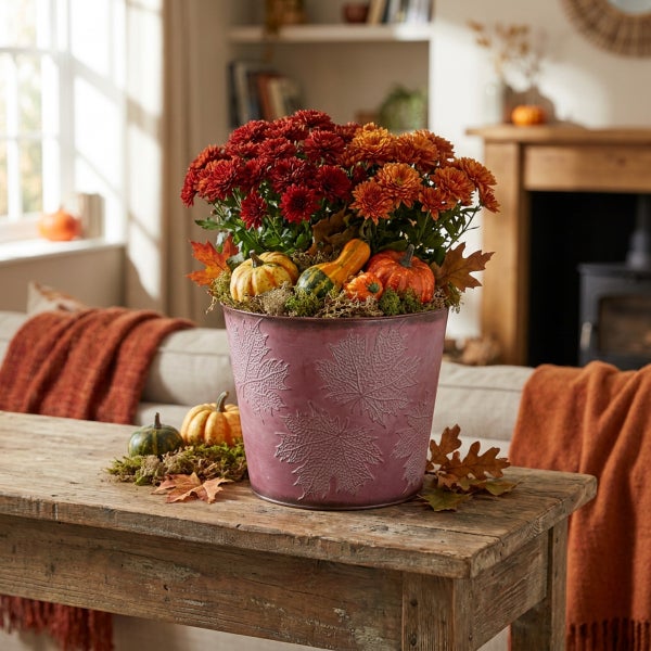 Herbstliche Blumendekoration mit Chrysanthemen und kleinen Zierkürbissen in einem Metalltopf mit Blattprägung auf einem Holztisch.