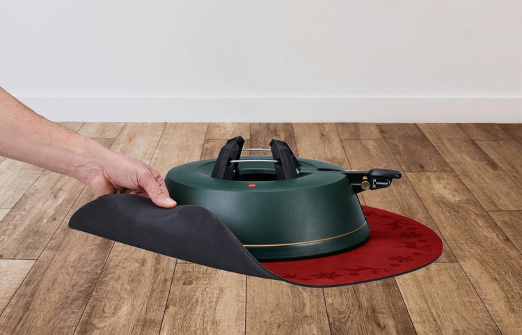 Grüner Krinner Christbaumständer auf einer runden, roten Bodenschutzmatte mit Schneeflockenmuster auf einem Holzboden.