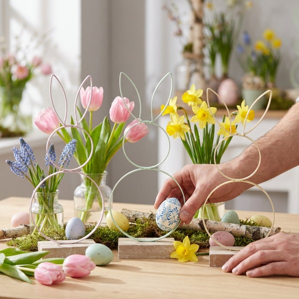 Drei Metallhasen als Osterdekoration auf Holzsockeln mit Frühlingsblumen und Ostereiern auf einem Holztisch dekoriert