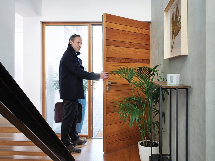 Mann öffnet eine hölzerne Haustür mit waagerechten Holzlatten in einem hellen Flur mit Smart Home Display und Zimmerpflanze.