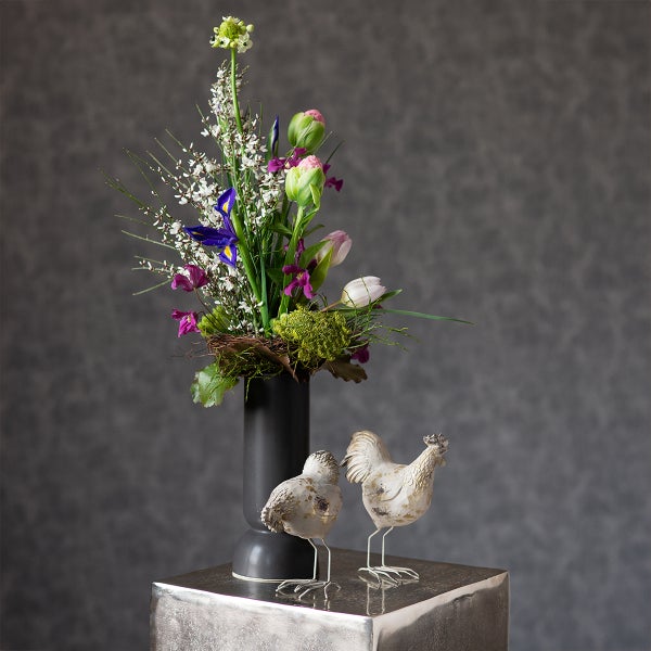 Moderner Blumenstrauß mit Tulpen und Iris in einer dunklen Vase, daneben zwei dekorative Hühnerfiguren auf einem silbernen Sockel.